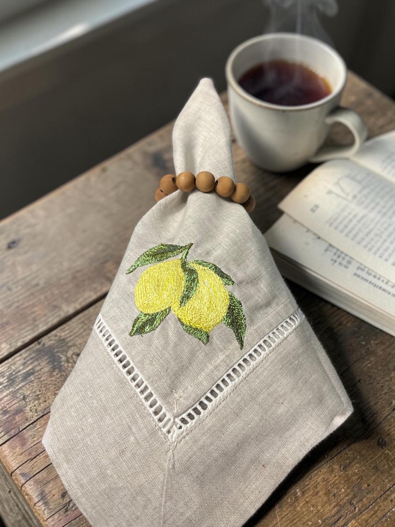 May include: A folded beige linen napkin with an embroidered lemon design, held by a wooden bead napkin ring. A cup of tea and an open book are in the background. The napkin features a decorative white border.