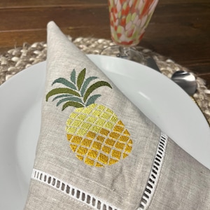 May include: A folded linen napkin with an embroidered pineapple design. The pineapple is yellow and green, with a detailed pattern. The napkin has a white, cut-out border and is placed on a white plate. A colorful glass is in the background.