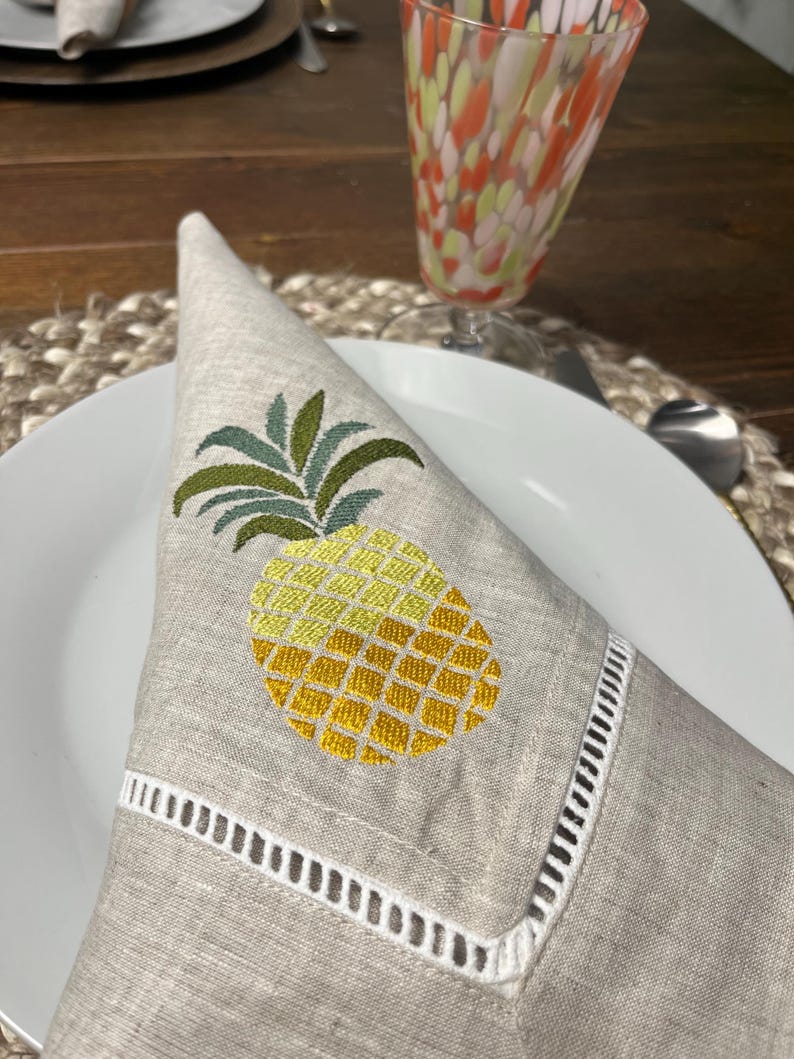 May include: A folded linen napkin with an embroidered pineapple design. The pineapple is yellow and green, with a detailed pattern. The napkin has a white, cut-out border and is placed on a white plate. A colorful glass is in the background.