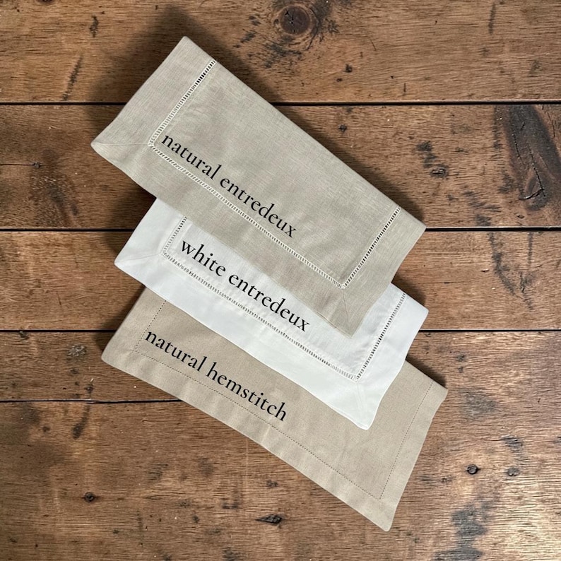 May include: Three linen napkins in neutral shades, displayed on a wooden surface. The napkins are in beige and white, featuring embroidered details and text reading "natural entredeux", "white entredeux", and "natural hemstitch".