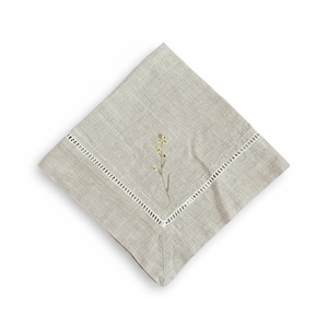 May include: A square, pale grey linen napkin with an embroidered floral design. The napkin has a white, cut-out border. The embroidered flower has yellow and green details, adding a touch of elegance. Ideal for a formal or casual dining setting.