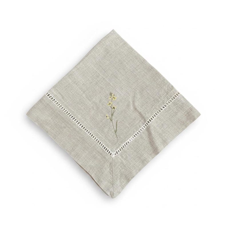May include: A square, pale grey linen napkin with an embroidered floral design. The napkin has a white, cut-out border. The embroidered flower has yellow and green details, adding a touch of elegance. Ideal for a formal or casual dining setting.