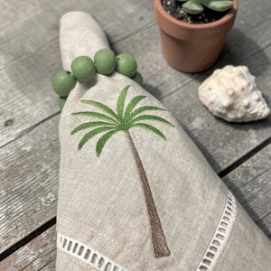 Pode incluir: Um guardanapo de linho bege com um desenho de palmeira bordada, preso por um porta-guardanapo de contas verdes. O guardanapo tem uma borda recortada branca. Uma pequena suculenta num vaso de terracota e uma concha est&atilde;o no fundo.