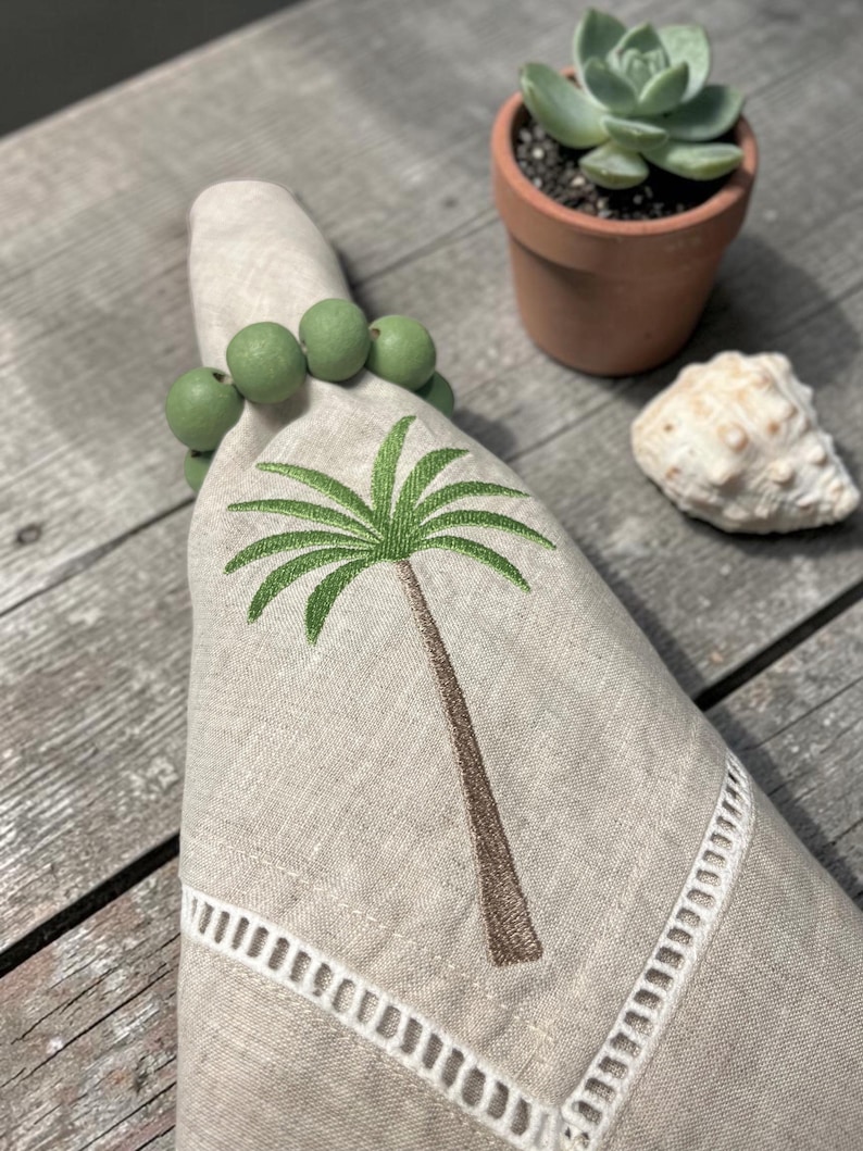 Pode incluir: Um guardanapo de linho bege com um desenho de palmeira bordada, preso por um porta-guardanapo de contas verdes. O guardanapo tem uma borda recortada branca. Uma pequena suculenta num vaso de terracota e uma concha est&atilde;o no fundo.