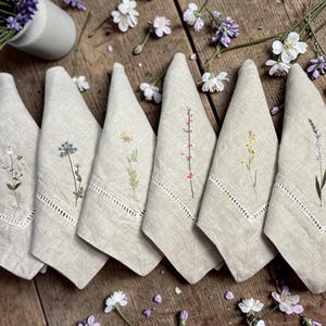 May include: Six beige linen napkins, each embroidered with a different wildflower design in green, pink, and yellow. The napkins have a decorative cutwork border. The napkins are arranged on a wooden surface.