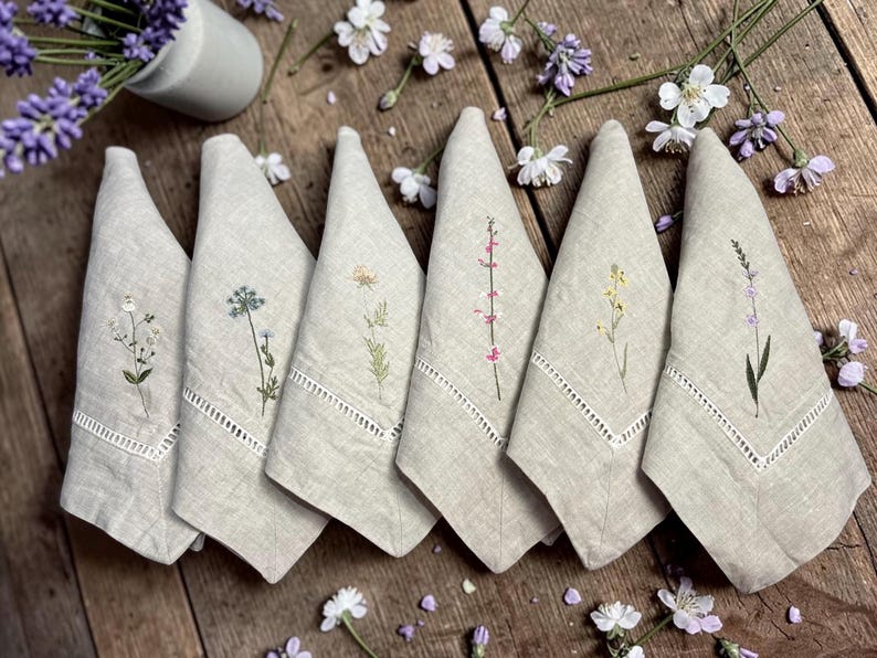 May include: Six beige linen napkins, each embroidered with a different wildflower design in green, pink, and yellow. The napkins have a decorative cutwork border. The napkins are arranged on a wooden surface.