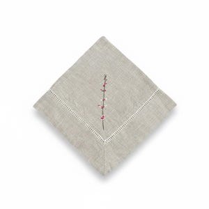 May include: A square, pale grey linen napkin with a white, stitched border. A delicate embroidered floral stem with pink and white blossoms is centered on the napkin. The napkin is folded in a diamond shape.