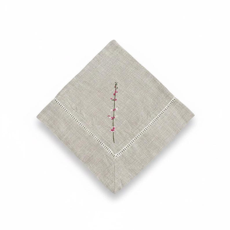 May include: A square, pale grey linen napkin with a white, stitched border. A delicate embroidered floral stem with pink and white blossoms is centered on the napkin. The napkin is folded in a diamond shape.