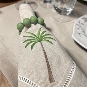 Pode incluir: Um guardanapo de linho com um bordado de palmeira e um porta-guardanapo de contas verdes. O guardanapo tem uma borda recortada branca. A palmeira &eacute; marrom e verde. O porta-guardanapo &eacute; feito de contas verdes.