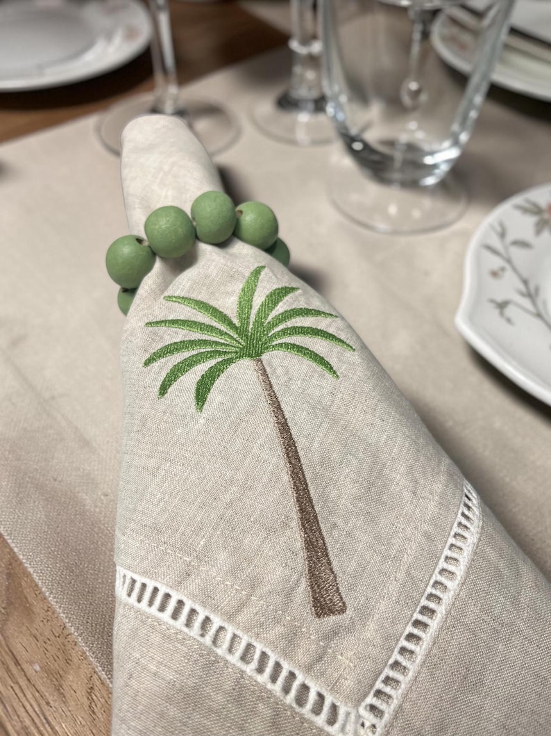 Pode incluir: Um guardanapo de linho com um bordado de palmeira e um porta-guardanapo de contas verdes. O guardanapo tem uma borda recortada branca. A palmeira &eacute; marrom e verde. O porta-guardanapo &eacute; feito de contas verdes.