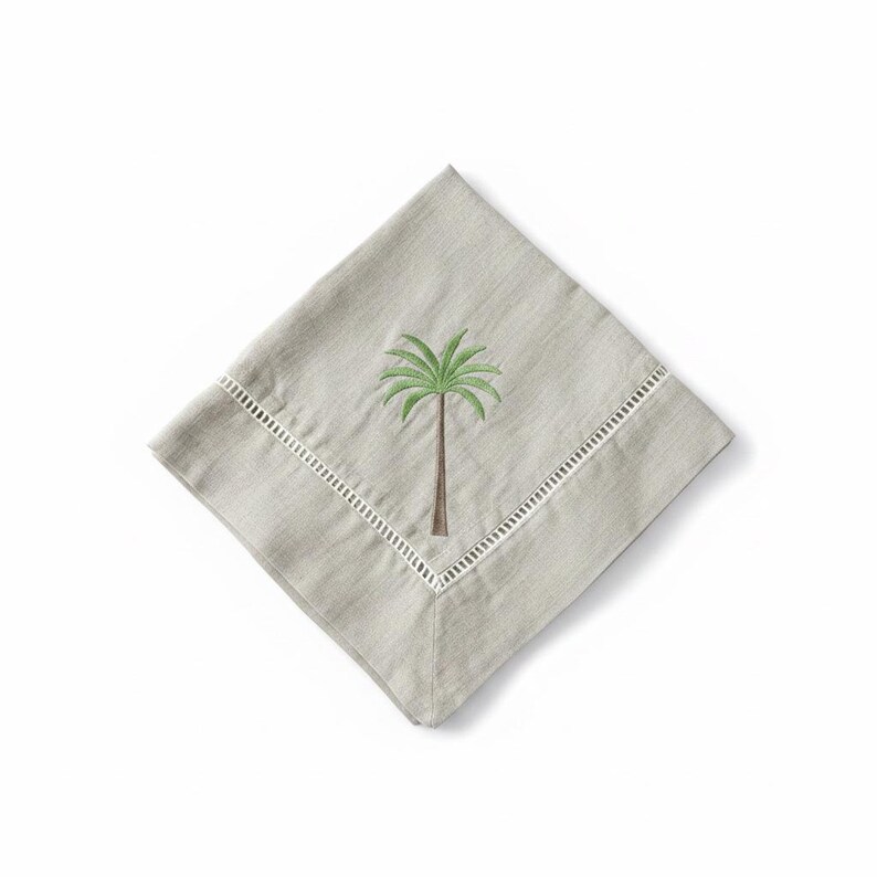 Pode incluir: Uma guardanapo quadrado cinza claro com uma borda vazada branca. O guardanapo apresenta um bordado de palmeira em verde e marrom. O guardanapo &eacute; dobrado em forma de diamante sobre um fundo branco.