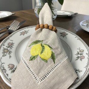 May include: A linen napkin with embroidered lemons and green leaves, held by a wooden bead napkin ring, rests on a floral patterned plate. The napkin has a cut-out border. A fork and knife are visible.