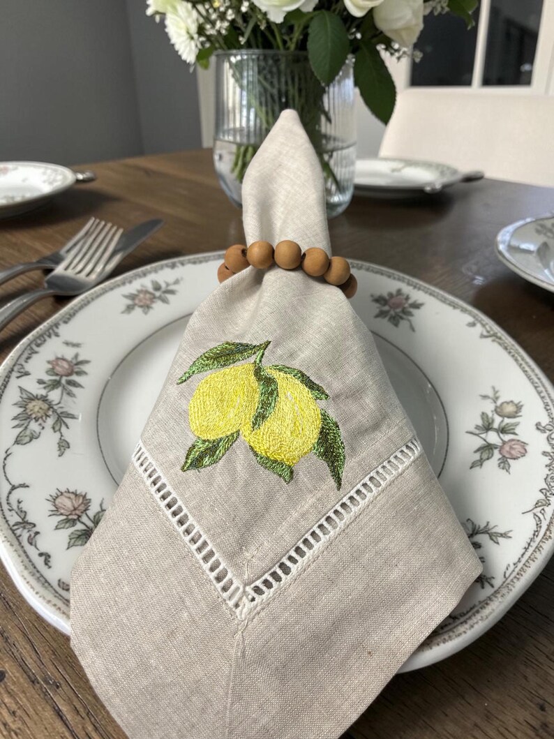 May include: A linen napkin with embroidered lemons and green leaves, held by a wooden bead napkin ring, rests on a floral patterned plate. The napkin has a cut-out border. A fork and knife are visible.