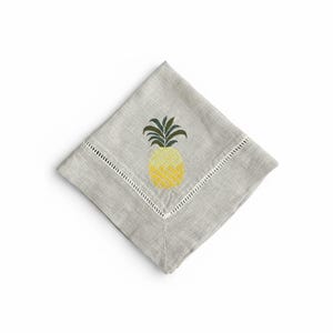 May include: A gray linen napkin with a yellow and green embroidered pineapple design. The napkin has a white stitched border and is folded in a diamond shape. The pineapple is centered on the napkin.