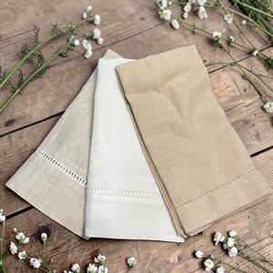 May include: Three folded linen napkins in shades of beige and white are arranged on a wooden surface. The napkins have decorative stitching and cut-out details along the edges. White flowers and greenery are scattered around the napkins.