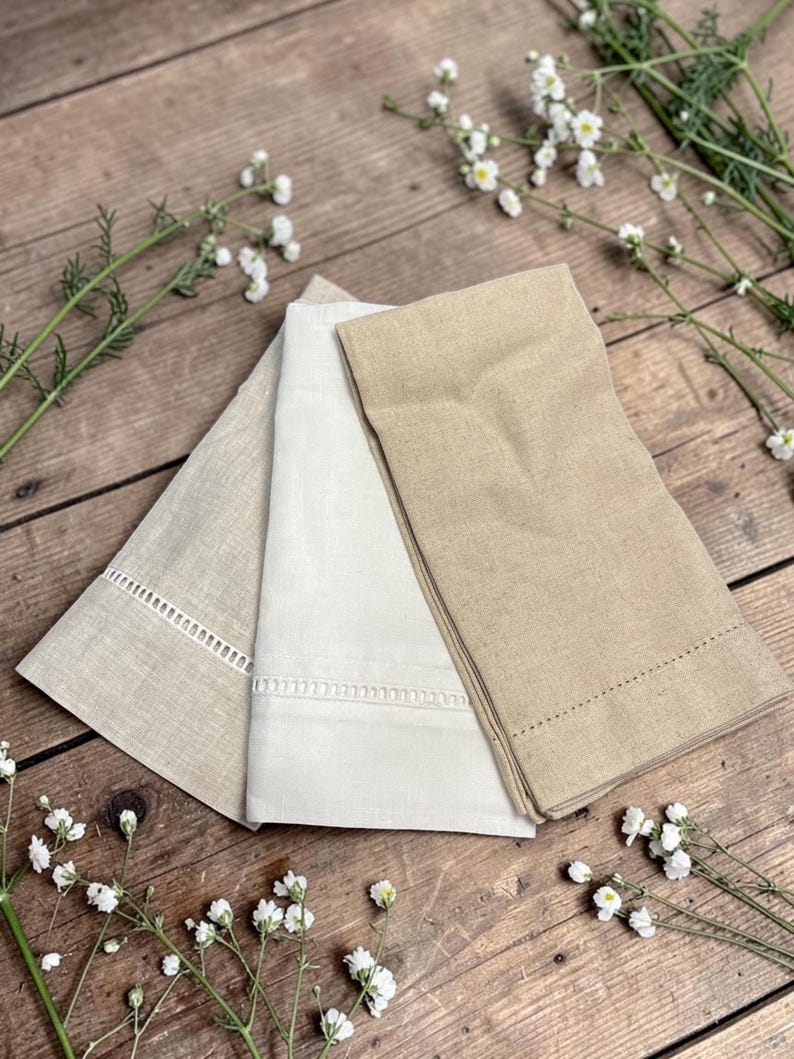 May include: Three folded linen napkins in shades of beige and white are arranged on a wooden surface. The napkins have decorative stitching and cut-out details along the edges. White flowers and greenery are scattered around the napkins.