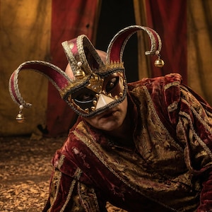 以下が含まれることがあります： 装飾的な道化師のマスクと赤と金の衣装を着た人。マスクは、赤いアクセントとぶら下がっているベルが付いた金と黒のデザインが特徴です。衣装は、演劇的または歴史的なテーマを連想させる、金色のトリムが付いた濃い赤色のベルベットです。