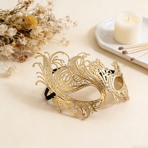 Puede incluir: Una máscara dorada para mascaradas con un diseño intrincado y calado. La máscara presenta patrones florales y de volutas, adornados con pequeños cristales transparentes. Una banda elástica negra asegura la máscara. Un accesorio decorativo.