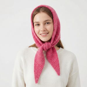 May include: A bright pink knitted headscarf, tied under the chin. The head covering is made of a textured material and is a solid, bright pink colour. The scarf is worn over a white jumper.