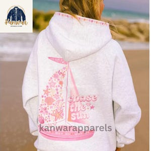 May include: A light gray hoodie with a pink sailboat design. The sailboat is decorated with seashells and starfish, and the text "Chase the Sun" is embroidered on the back. The hoodie also features the text "kanwarapparels".