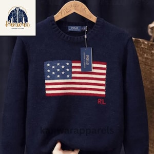 May include: Navy blue sweater featuring an American flag design on the front. The flag has white stars on a blue background and red and white stripes. The sweater has a crew neck and the brand name "Polo" is visible on a tag.