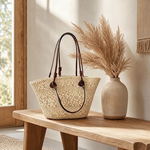 May include: A woven straw tote bag featuring a light beige natural texture and two thin dark brown leather shoulder straps attached with circular leather tabs. The bag has an open top and a structured, tapered shape with a detailed basket weave pattern.