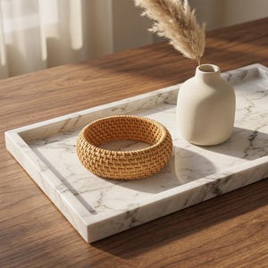 Puede incluir: Una bandeja rectangular de m&aacute;rmol blanco con vetas grises que presenta una cesta redonda de rat&aacute;n tejido y un peque&ntilde;o jarr&oacute;n de cer&aacute;mica beige mate con una pluma de hierba de la pampa seca. La bandeja tiene bordes elevados y un acabado liso.