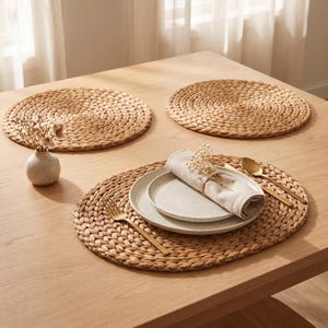 May include: Three round woven seagrass placemats arranged on a light wood table. One setting includes a speckled ceramic plate, a folded beige linen napkin with dried flowers, a gold fork, and a gold spoon. A small ceramic vase with dried flowers sits nearby.