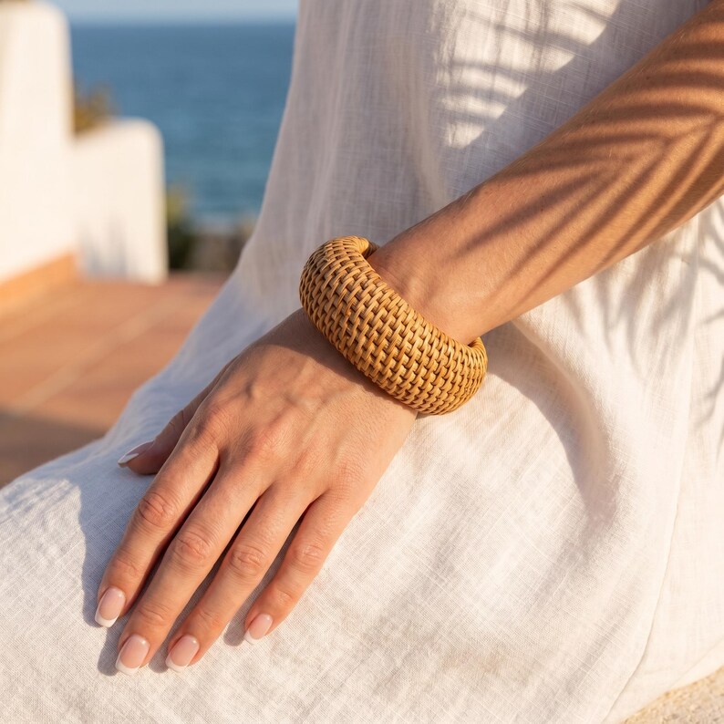 Puede incluir: Una pulsera tipo brazalete ancha y cil&iacute;ndrica hecha de material de rat&aacute;n tejido en color marr&oacute;n claro. El accesorio presenta un patr&oacute;n de tejido de cesta denso y uniforme en toda su superficie, creando una apariencia redondeada y texturizada.