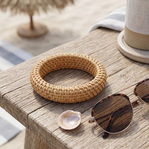 Puede incluir: Un brazalete redondo y grueso elaborado con material de rat&aacute;n tejido en color tostado claro. La pulsera presenta un patr&oacute;n de tejido de cesta apretado y consistente en toda su estructura circular, con un acabado natural, texturizado y marr&oacute;n claro.