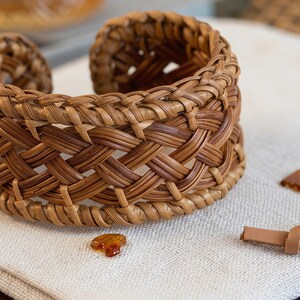 Puede incluir: Una pulsera de brazalete ancha hecha de fibras naturales tejidas de color marr&oacute;n en un patr&oacute;n de celos&iacute;a complejo. El dise&ntilde;o presenta un borde trenzado grueso a lo largo de los bordes superior e inferior, creando una estructura abierta y resistente.