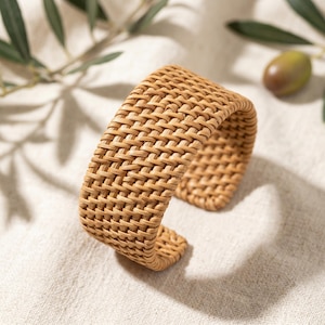 May include: A wide cuff bracelet constructed from light brown woven rattan material. The accessory features a continuous, intricate basket-weave pattern throughout its entire surface, forming a rigid, open-ended circular structure for the wrist.