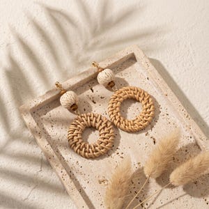 Pode incluir: Um par de brincos de rattan tecido com um design circular. Os brincos são bege e são exibidos em uma bandeja de pedra. Cada brinco apresenta uma pequena conta redonda de cor creme e uma conta de madeira no topo.