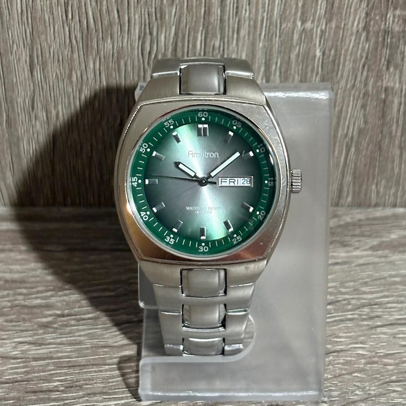 May include: A silver Armitron watch with a green face and bezel. The watch shows the day and date, with the brand name and the word "QUARTZ" visible. The watch is displayed on a clear stand. The watch has a silver strap.
