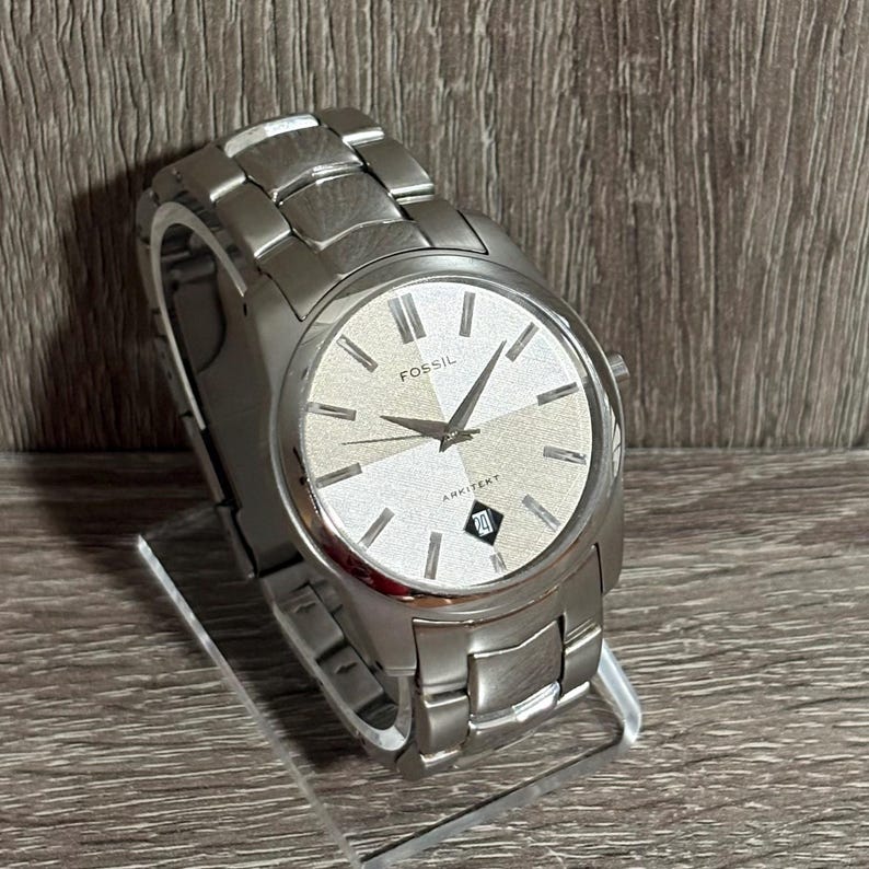 Puede incluir: Un reloj Fossil Arkitekt plateado con una correa plateada. La esfera del reloj es plateada con manecillas y marcadores negros. La fecha se muestra a las 6 en punto. El reloj se exhibe sobre un soporte transparente.