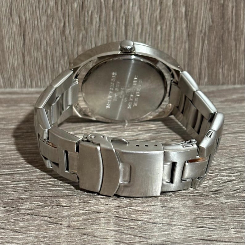 May include: A stainless steel wristwatch with a silver strap and clasp. The watch face is not visible. The back of the watch has engraved text. The watch is resting on a wooden surface.