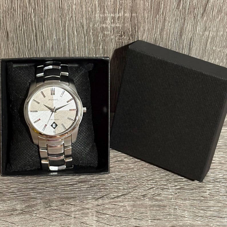 Puede incluir: Un reloj Fossil plateado con esfera clara y agujas negras, presentado en una caja negra. El reloj tiene una correa plateada con detalles negros. La esfera del reloj tiene el logotipo de Fossil y la palabra "water resistant".