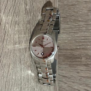 Puede incluir: Un reloj Fossil plateado con esfera rosa claro y correa plateada. La esfera muestra el logotipo de Fossil y la palabra "AMBITION" debajo. El reloj est&aacute; sobre una superficie de madera.