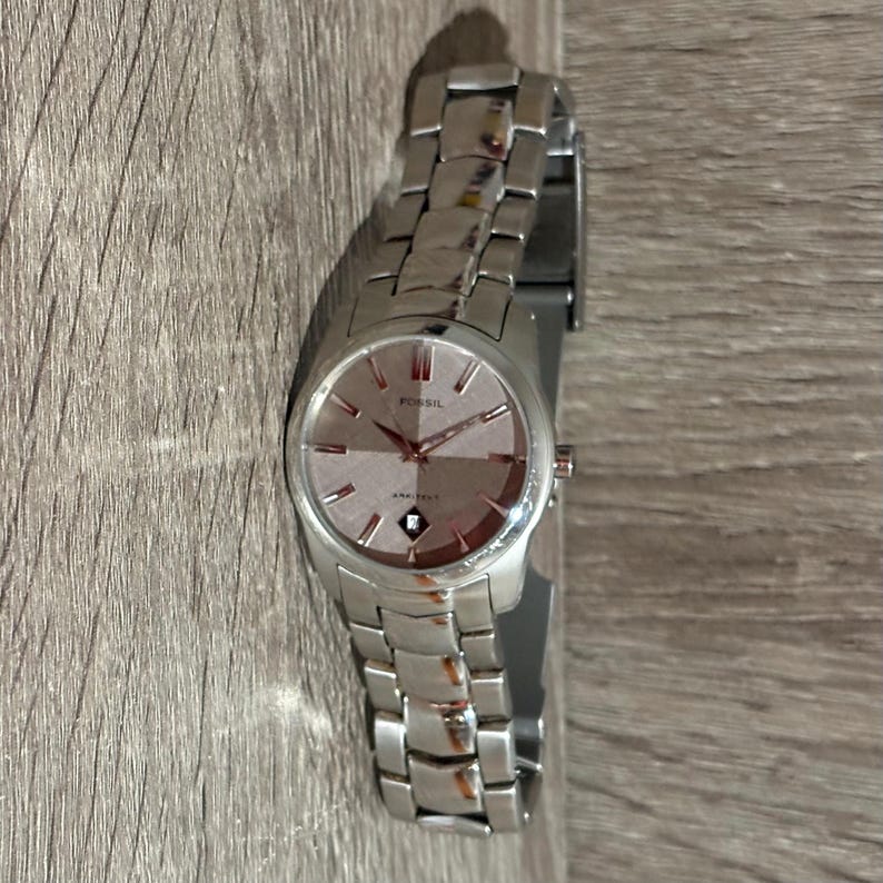 Puede incluir: Un reloj Fossil plateado con esfera rosa claro y correa plateada. La esfera muestra el logotipo de Fossil y la palabra "AMBITION" debajo. El reloj est&aacute; sobre una superficie de madera.