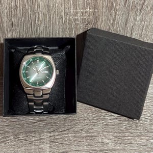 May include: A silver wristwatch with a green face and a black strap is presented in a black box. The watch face has white markings and the day of the week is visible. The watch is on a black cushion.