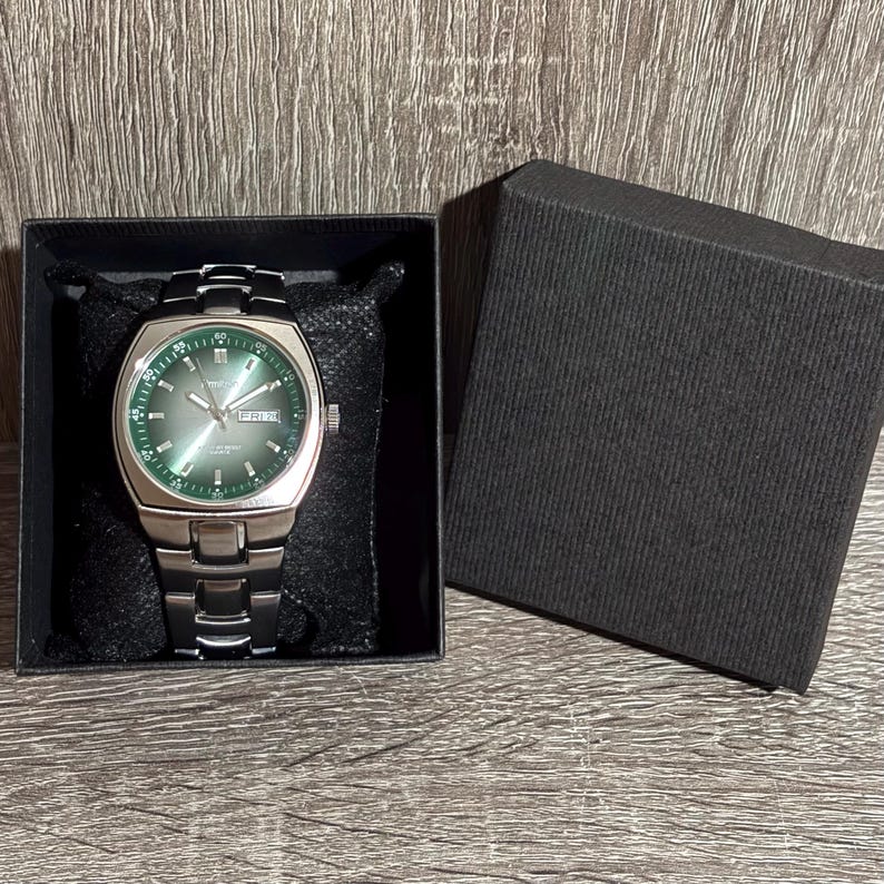 May include: A silver wristwatch with a green face and a black strap is presented in a black box. The watch face has white markings and the day of the week is visible. The watch is on a black cushion.