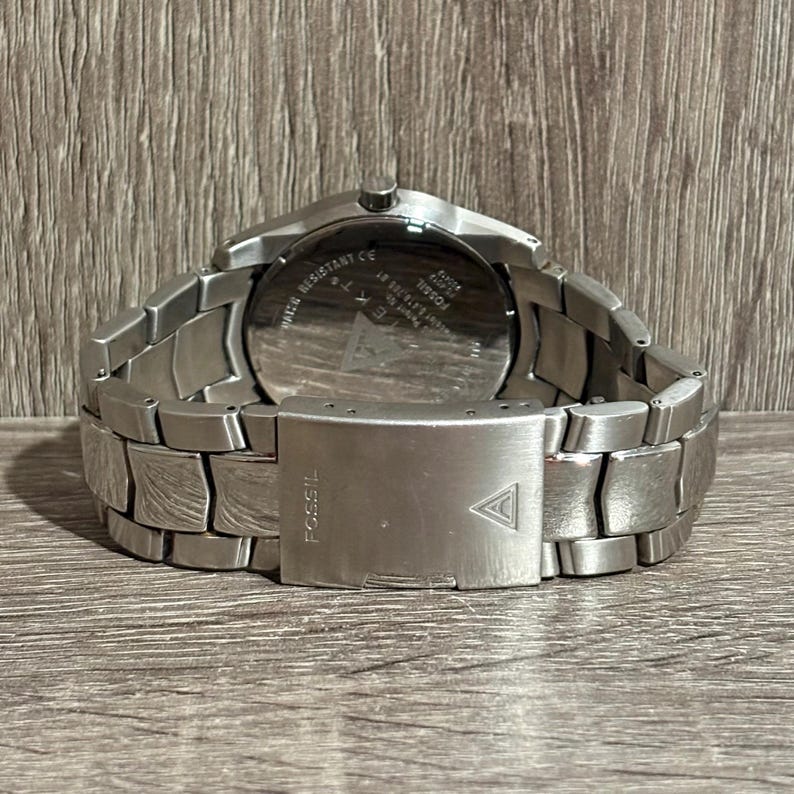 Puede incluir: Un reloj Fossil de acero inoxidable con una correa plateada. El reloj tiene una esfera redonda y un cierre con el logotipo de Fossil. La parte trasera del reloj tiene el texto "Water Resistant CE".