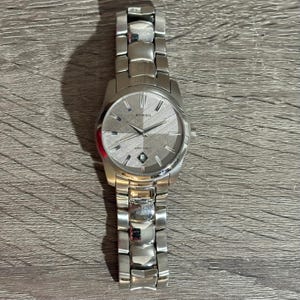Puede incluir: Un reloj Fossil plateado con una esfera gris texturizada y una pantalla de fecha. El reloj tiene una correa de acero inoxidable y una esfera redonda. El reloj est&aacute; sobre una superficie de madera.