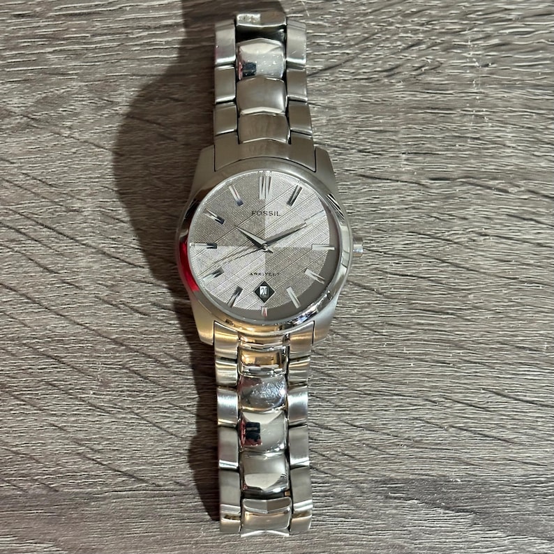 Puede incluir: Un reloj Fossil plateado con una esfera gris texturizada y una pantalla de fecha. El reloj tiene una correa de acero inoxidable y una esfera redonda. El reloj est&aacute; sobre una superficie de madera.