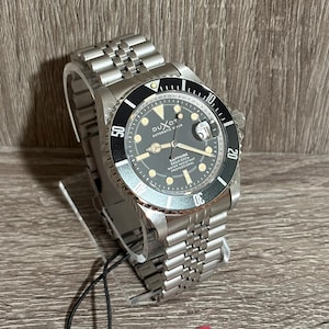 May include: A silver-toned wristwatch with a black rotating bezel and a dark gray face. The watch features the brand name "DUXOT" and the text "AUTOMATIC DIVER". The watch band is made of silver-toned metal links.