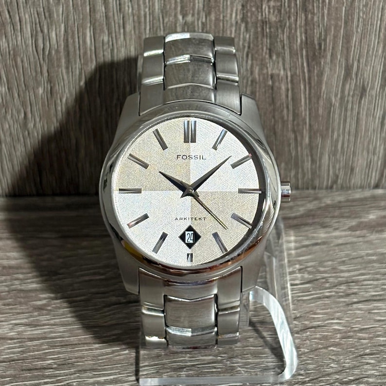 Puede incluir: Un reloj Fossil Arkitekt plateado con esfera plateada texturizada, manecillas negras y visualizaci&oacute;n de la fecha a las 6. El reloj tiene una correa de acero inoxidable y se muestra sobre un soporte transparente.