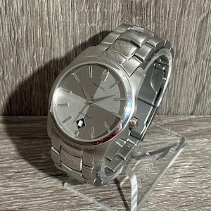 Puede incluir: Un reloj Fossil Arkitekt plateado con esfera gris de dos tonos. El reloj tiene una correa de acero inoxidable y una pantalla de fecha a las 6 en punto. El reloj se exhibe sobre un soporte transparente.