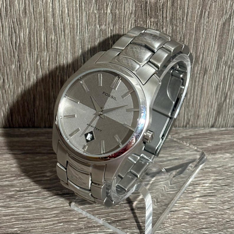 Puede incluir: Un reloj Fossil Arkitekt plateado con esfera gris de dos tonos. El reloj tiene una correa de acero inoxidable y una pantalla de fecha a las 6 en punto. El reloj se exhibe sobre un soporte transparente.