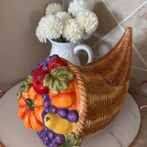 Puede incluir: Una cornucopia de cerámica rebosante de coloridas frutas y verduras esculpidas. La cesta en forma de cuerno es marrón con una textura tejida. El arreglo incluye calabazas, uvas y tomates. La cornucopia se encuentra sobre una bandeja de mármol blanco.