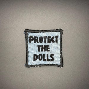 Peut inclure: Écusson carré avec le texte "PROTECT THE DOLLS" en lettres noires. Le patch a un fond en denim et une bordure noire cousue. Le patch est centré sur une surface grise.